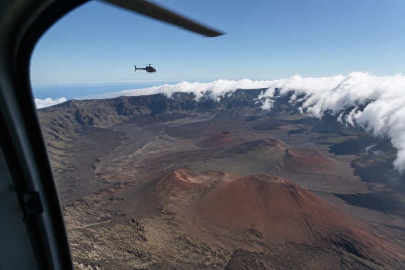 View from a helicopter window of reddish volcanic cinder cones and craters with a distant helicopter amid clouds.