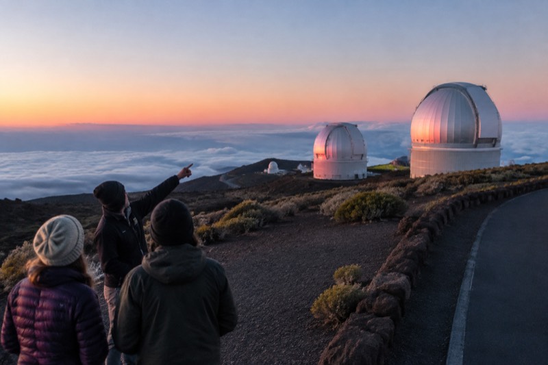 Four people at Haleakala summit view white observatory domes on the mountain at sunset above a sea of clouds.