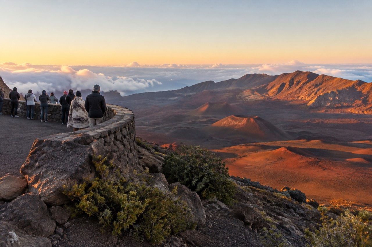 Visitors on a stone wall overlook above reddish volcanic slopes and clouds under a golden sunset sky at Haleakala summit.