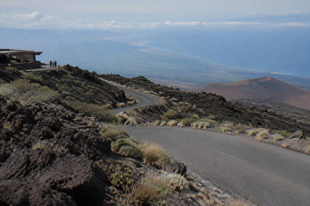 Winding paved mountain road descending through lava rock landscape with distant coastline