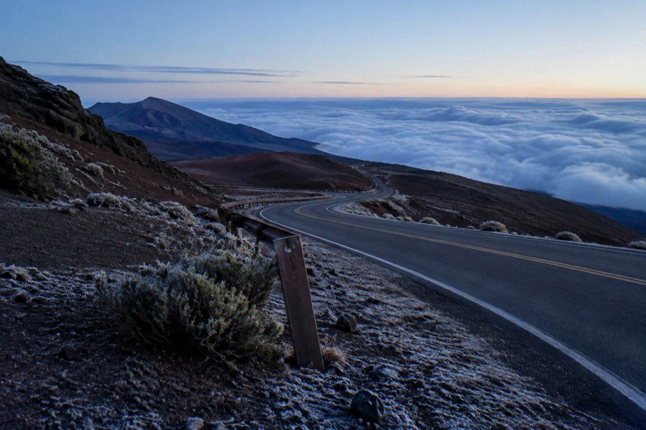 Empty crater road curving downhill across dark lava slopes below clouds