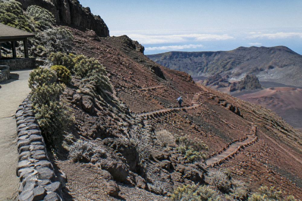 A footpath of loose dark cinders switchbacking down a red and gray crater wall