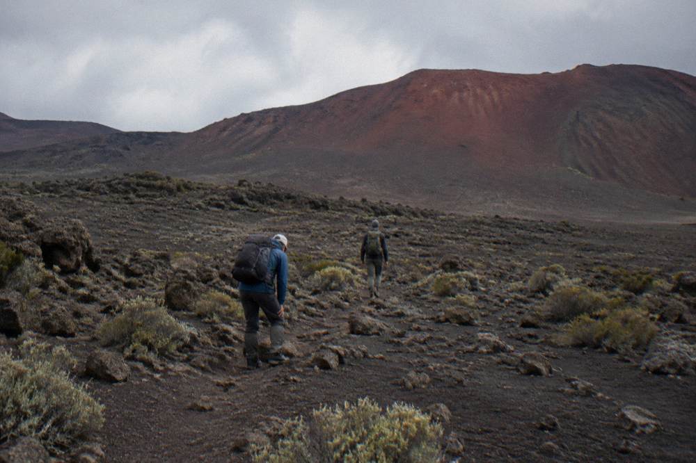 Hikers on a narrow dirt path crossing a plain of dark volcanic rock below a red rim