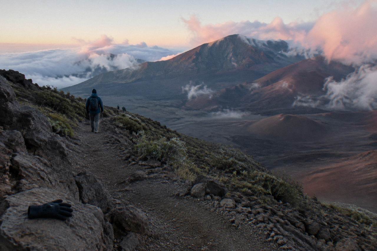 A long trail of dark volcanic cinders descending into a wide crater below morning clouds