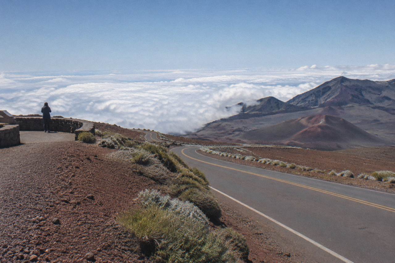 A quiet two-lane road curving down the open slopes of Haleakala above a layer of clouds