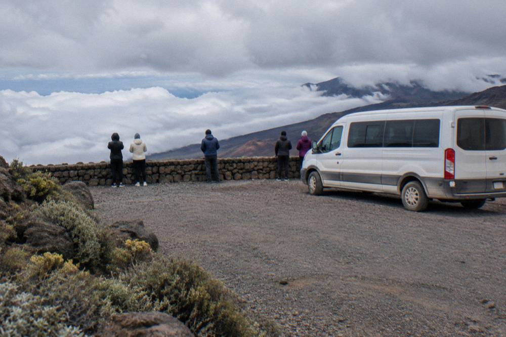 A parked tour van at a crater overlook with visitors in winter jackets looking out over clouds