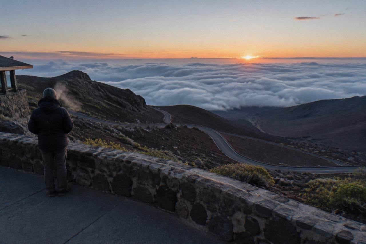 A volcanic summit ridge rising above a sea of clouds at dawn, with a winding road visible far below